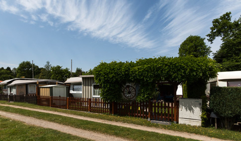 Eine Reihe kleiner Mobilheime mit eingezäunten Gärten und grüner Vegetation entlang einer unbefestigten Straße unter einem blauen Himmel mit vereinzelten Wolken.