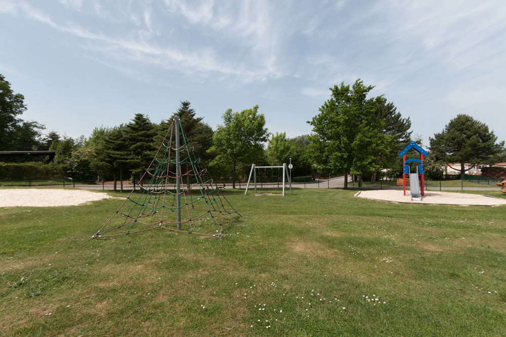 Ein Spielplatz mit einem Kletternetz, Schaukeln und einer Spielhausrutsche auf einer von Bäumen umgebenen Wiese unter einem teilweise bewölkten Himmel.