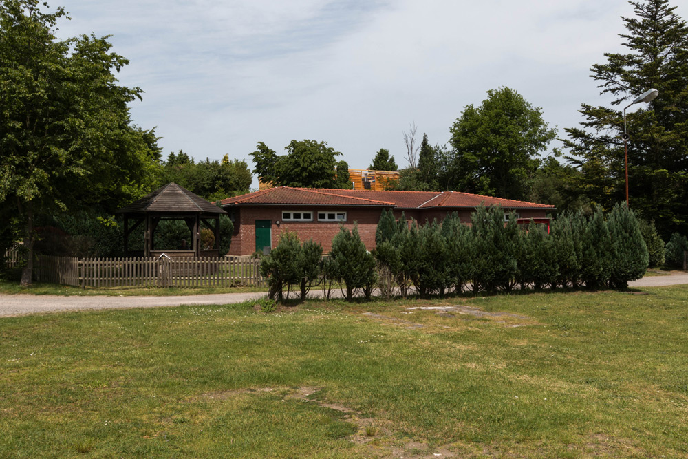 Einstöckiges Backsteingebäude mit rotem Ziegeldach, grüner Tür und kleinen Fenstern, umgeben von Bäumen, Sträuchern und einer Rasenfläche; links davon ein Holzpavillon.