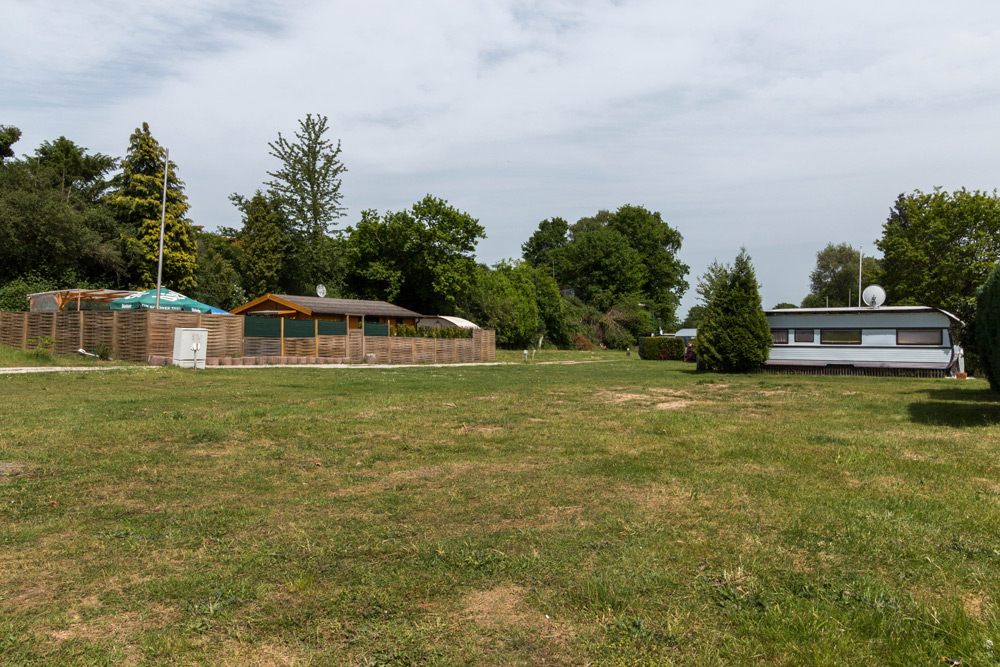 Eine grasbewachsene Freifläche mit einem Holzzaun, einigen Gebäuden und einem weißen Wohnwagen unter einem teilweise bewölkten Himmel, umgeben von Bäumen.
