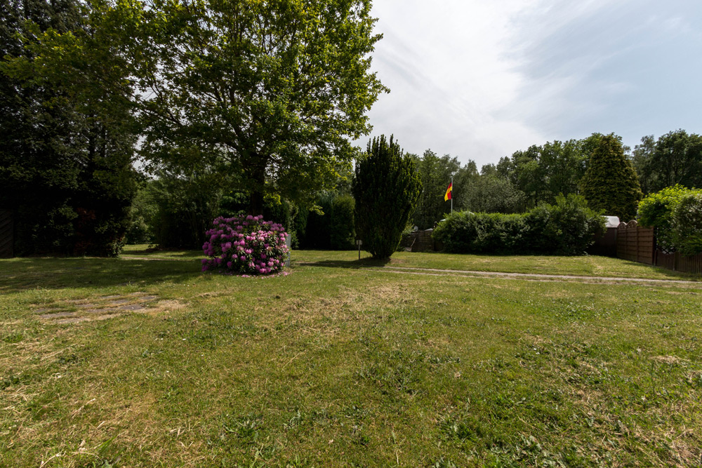Ein grasbewachsener Hof mit einem blühenden violetten Strauch, mehreren Bäumen und einer Fahne in der Ferne unter einem teilweise bewölkten Himmel.
