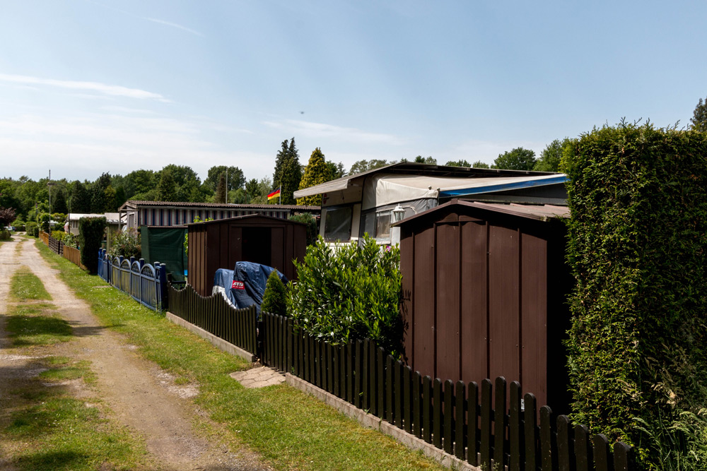 Eine Reihe von kleinen eingezäunten Höfen und Schuppen neben einem Kiesweg in einem Wohn- oder Kleingartengebiet mit Grünflächen und Bäumen im Hintergrund.
