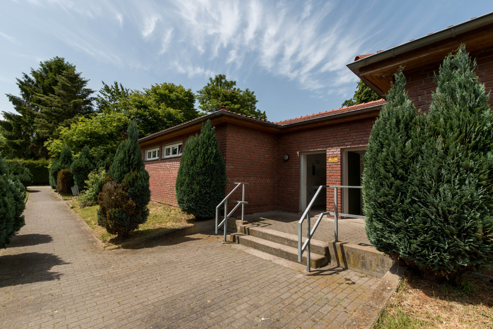 Ein Backsteingebäude mit rotem Dach, kleinen Fenstern und einem Eingang mit Metallgeländer, umgeben von Bäumen und Grünpflanzen an einem sonnigen Tag.