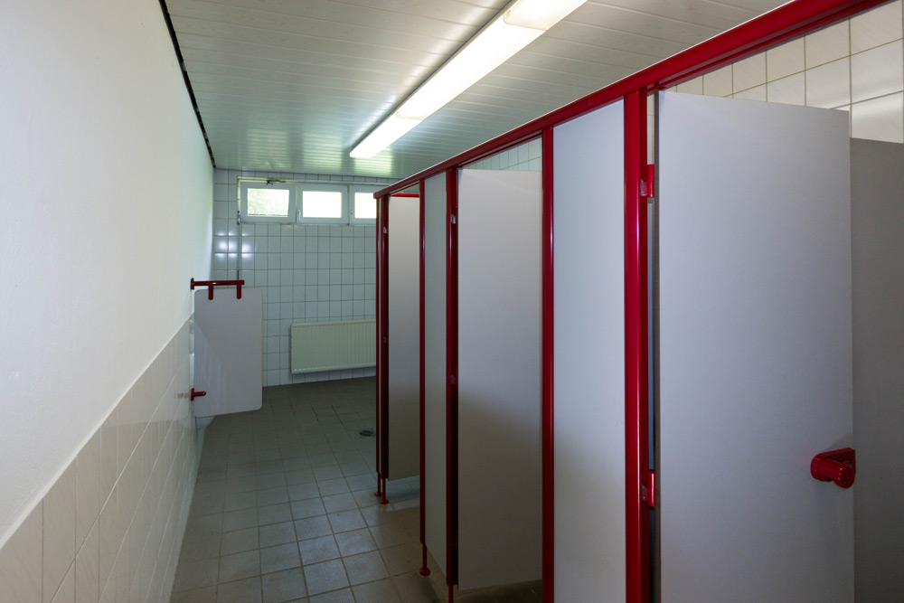 Eine öffentliche Toilette mit rot umrahmten Kabinentüren, gefliesten Wänden und Böden, einem Heizkörper und einer fluoreszierenden Deckenleuchte.