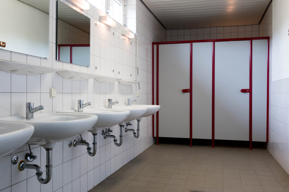 Reihe weißer Waschbecken mit Spiegeln an der linken Seite einer gefliesten öffentlichen Toilette; im Hintergrund sind rot umrahmte Kabinen mit geschlossenen Türen zu sehen.