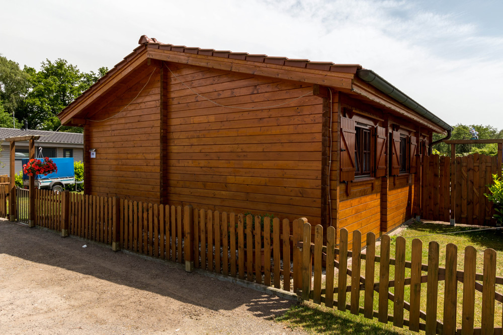 Eine kleine Holzhütte mit verschlossenen Fenstern steht an einem sonnigen Tag hinter einem Lattenzaun, und ein blaues Auto ist in der Nähe geparkt.