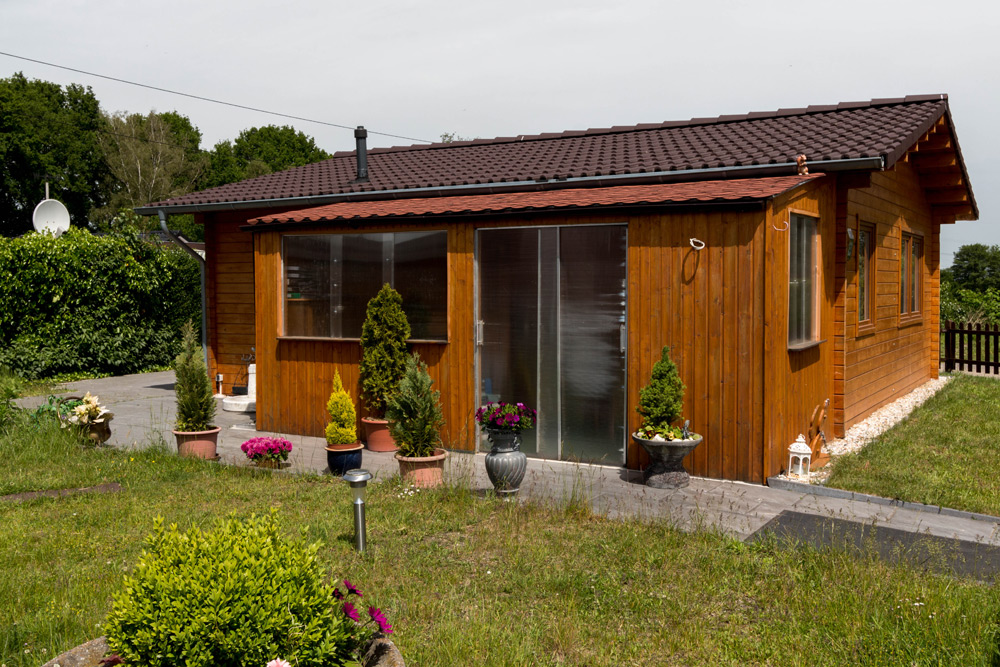 Ein kleines Holzhaus mit einem braunen Ziegeldach, großen Glasschiebetüren und Topfpflanzen auf der Terrasse in einem grasbewachsenen Hof.