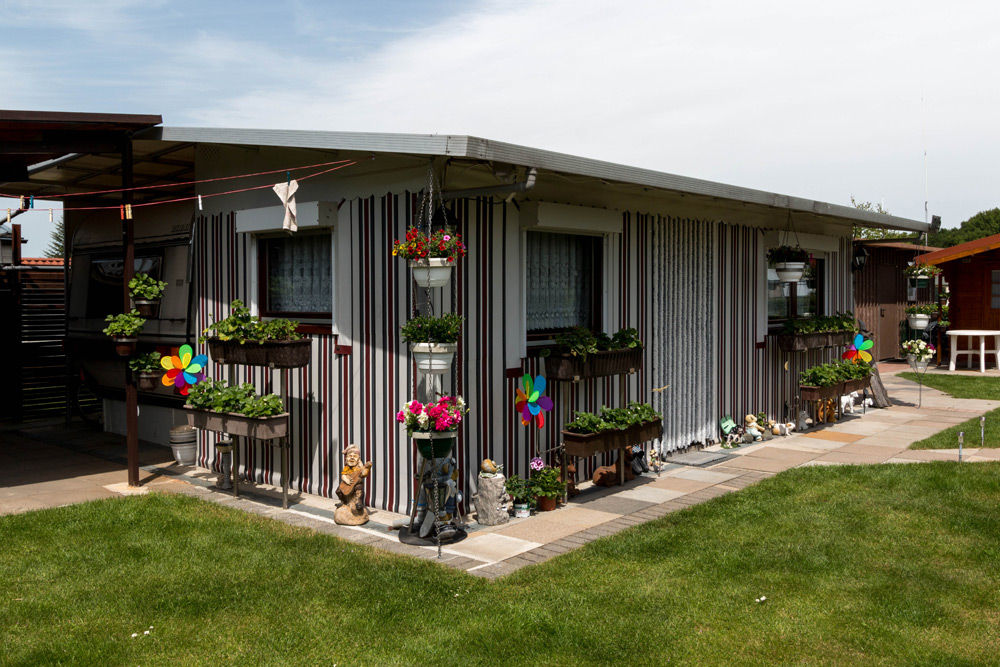 Einstöckiges Haus mit gestreiftem Äußeren, Blumenkästen, Gartendekorationen und Windrädern, umgeben von einem grünen Rasen unter einem teilweise bewölkten Himmel.