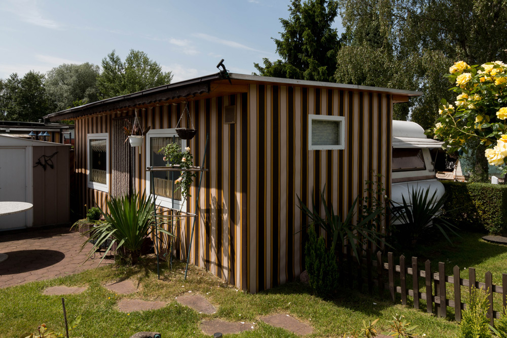Kleines, einstöckiges Gebäude mit vertikalen braunen und gelben Streifen, umgeben von Pflanzen und Gras, neben einem weißen Schuppen und einem Wohnmobil.