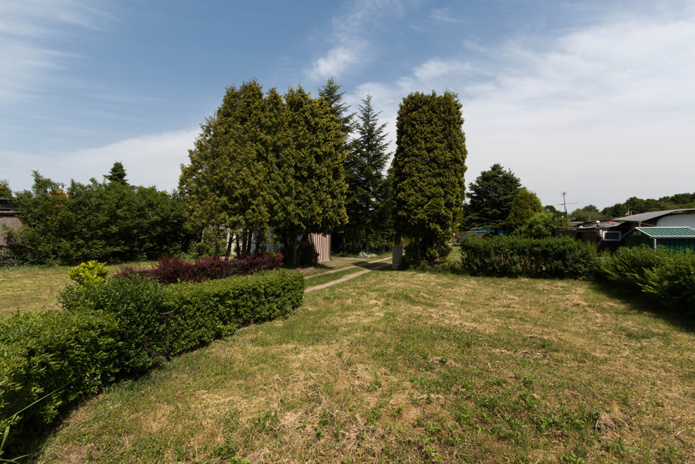 Ein grasbewachsener Hof mit beschnittenen Büschen, hohen Bäumen und einem Weg, der zu einem kleinen Schuppen unter einem blauen Himmel mit vereinzelten Wolken führt.