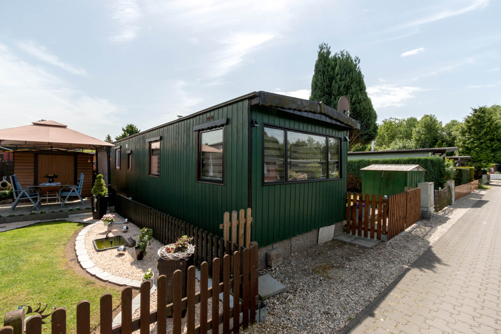 Ein kleines, dunkelgrünes Wohnmobil mit großen Fenstern, umgeben von einem Garten, einem Lattenzaun und einem gepflasterten Spazierweg unter einem teilweise bewölkten Himmel.