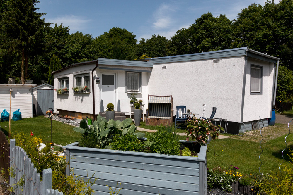Ein kleines weißes einstöckiges Haus mit blauen Zierleisten, einem Vorgarten mit Blumen und Gemüse, Sitzgelegenheiten im Freien und Bäumen im Hintergrund unter einem klaren Himmel.