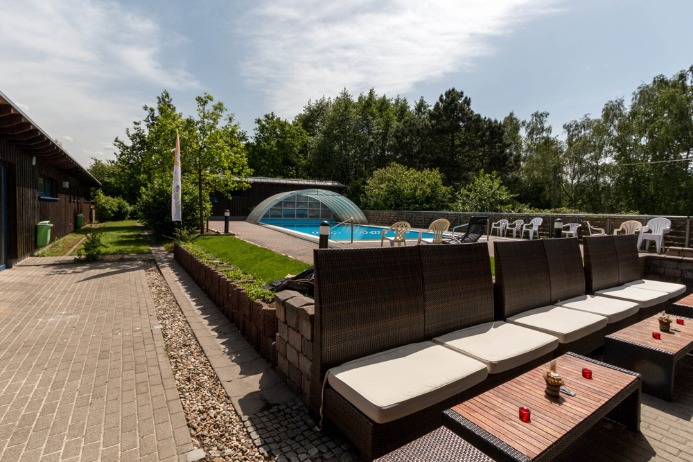 Sitzbereich im Freien mit gepolsterten Bänken und Tischen in der Nähe des Swimmingpools, der teilweise von einer transparenten Kuppel überdacht ist, umgeben von Bäumen und Grün.