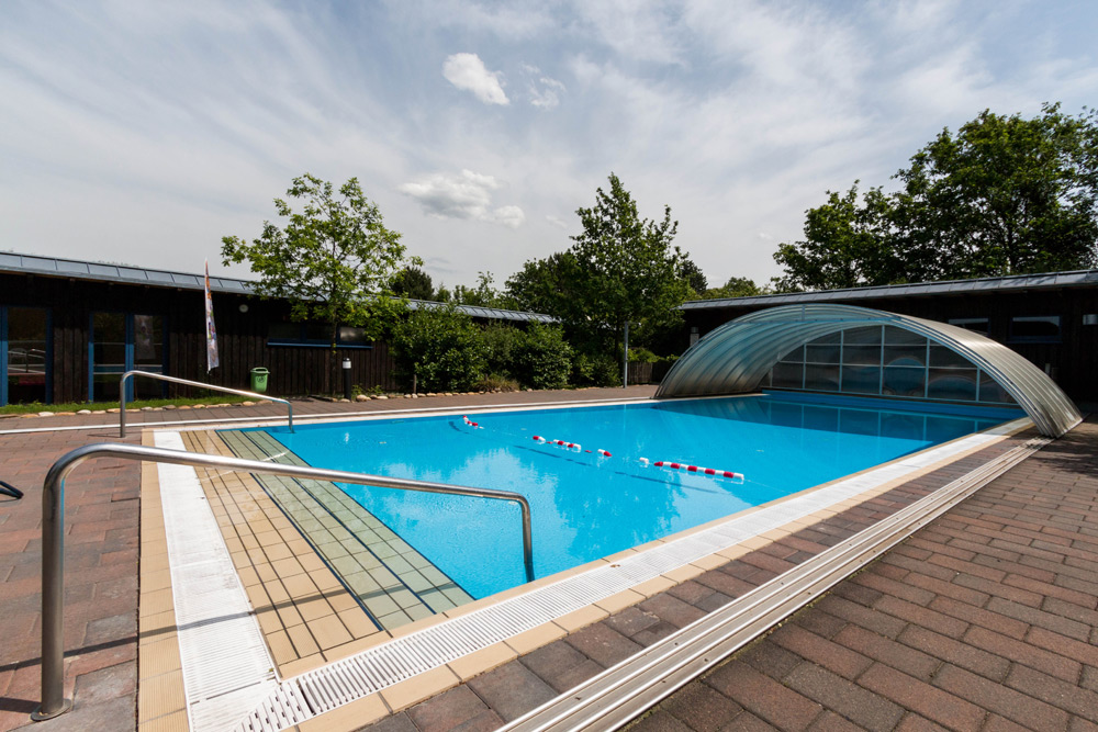 Außenschwimmbad mit teilweise transparenter Abdeckung, Bahntrennern, Handläufen und umlaufender Pflasterung, unter teilweise bewölktem Himmel.