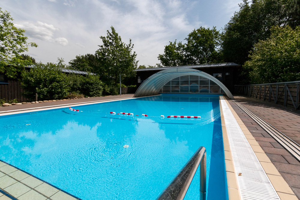 Freibad mit klarem, blauem Wasser, Sicherheitsseilen und einem teilweise überdachten Bereich; umgeben von Bäumen und gepflasterten Wegen unter einem teilweise bewölkten Himmel.
