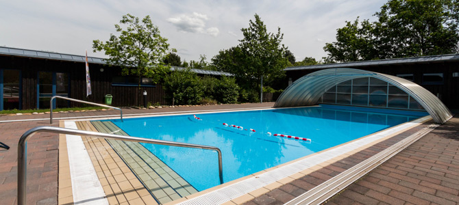 Freibad mit klarem, blauem Wasser, Bahntrennern, Metallgeländern, einer teilweise einziehbaren Abdeckung und Bäumen und Gebäuden im Hintergrund.