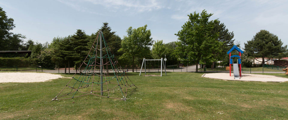 Ein Spielplatz mit einem Seilklettergerüst, einer Schaukel und einer Rutsche befindet sich auf einer Wiese, die an einem klaren Tag von Bäumen umgeben ist.