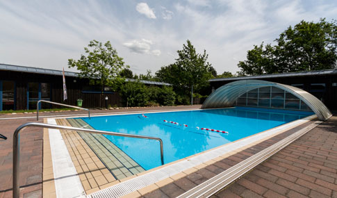 Außenschwimmbad mit teilweise versenkbarer Abdeckung, Handläufen und Bahnmarkierungen; an sonnigen Tagen umgeben von Bäumen und einer gepflasterten Fläche.