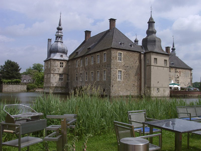 Ein steinernes Schloss mit Türmen und einem Wassergraben, betrachtet von einem Sitzplatz im Freien mit Metalltischen und Stühlen im Vordergrund.